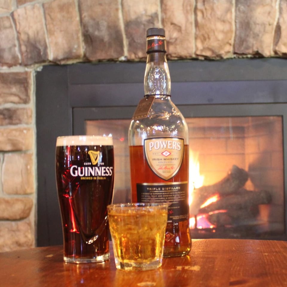 Guinness pint, whiskey shot, and Powers Irish Whiskey bottle on a table by a fireplace.