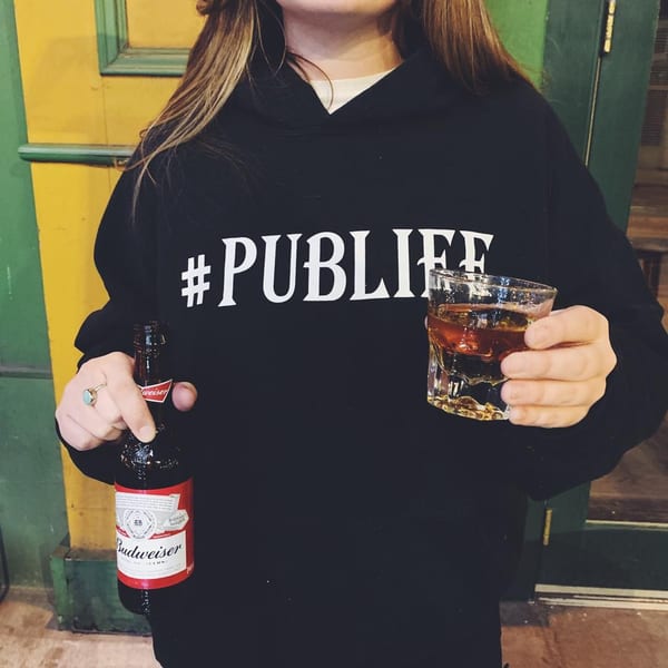 Person wearing a "#PUBLIFE" hoodie, holding a Budweiser bottle and a glass of whiskey.