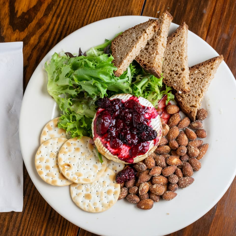 Baked brie with berry compote, crackers, almonds, mixed greens, and brown bread on a white plate.